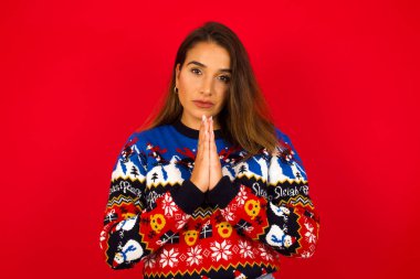 Genç, beyaz bir kadın, kırmızı duvara karşı Noel süveteri giyiyor. Avuçlarını önünde birbirine bastırıyor. Pişman görünüyor, af diliyor. Lütfen beni affet..