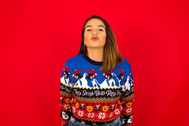 Genç, beyaz bir kadın, kırmızı duvara karşı Noel süveteri giyiyor, dudaklarını öpecek gibi tutuyor, mutlu bir ifade takınıyor, suratını buruşturuyor. Kapalı yerde dikiliyorum. Güzellik konsepti.