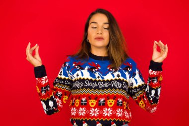 Kızıl duvara karşı Noel süveteri giyen genç, beyaz bir kadın yoga yapıyor, gözlerini kapalı tutuyor, Mudra jesti için parmaklarını tutuyor. Meditasyon, din ve ruhani uygulamalar.
