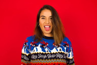 Vücut dili. Kırmızı duvara karşı Noel süveteri giyen, suratını asan, bir şeyden tiksindiğini gösteren, stresli, genç, beyaz bir kadın..