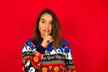 Genç, beyaz bir kadın, kırmızı duvara karşı Noel süveteri giyiyor, sessiz ol diyor. Sırrımı kimseye söyleme ya da yüksek sesle konuşma, lütfen.!