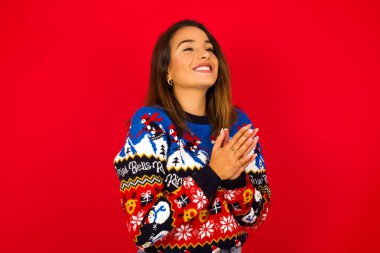 Genç, beyaz bir kadın, kırmızı duvara karşı Noel süveteri giyiyor, mutlu hissediyor, gülümsüyor ve alkışlıyor, alkışlarla tebrik ediyor..