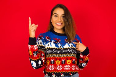 Genç, beyaz bir kadın, kırmızı duvara karşı Noel süveteri giyiyor ve diyor ki: Vay be, ne kadar heyecan verici, hayret verici bir ifade, bir şey ifade ediyor. Bir eli göğsünde ve diğer eliyle işaret ediyor..