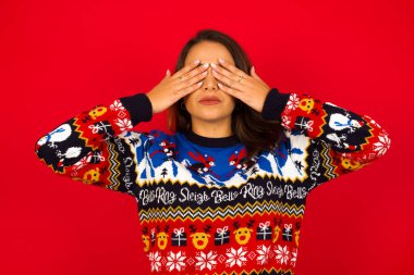 Genç, beyaz bir kadın, kırmızı duvara karşı Noel süveteri giyiyor, iki eliyle gözlerini kapatıyor, hiçbir şey görmek ya da utanmak istemiyor. İnsani duygular tepki verir.