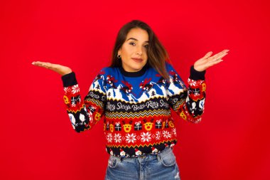 Şaşırmış ve cahil genç, beyaz bir kadın Noel süveterini kırmızı duvara dayamış, kollarını açmış, omuzlarını silkip: 