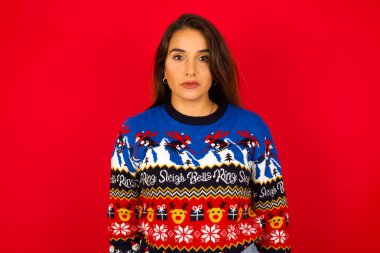 Kırmızı duvara karşı Noel süveteri giyen, kameraya bakıp bir şey düşünen neşeli, genç, beyaz bir kadın. İki kolunu da indir, tarafsız yüz ifadesi..