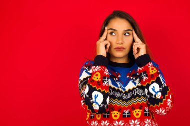 Genç, güzel, beyaz bir kadın, kırmızı duvara karşı Noel süveteri giyiyor, düşünceli bir ifadeyle, başka yere bakıyor, yüzünü çeviriyor, hoş bir şeyler düşünüyor..