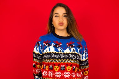 Genç, güzel, beyaz bir kadın Noel süveteri giyip kırmızı duvara yaslanmış, yanaklarını komik bir suratla şişiriyor. Havayla şişmiş ağız, çılgın bir ifade..