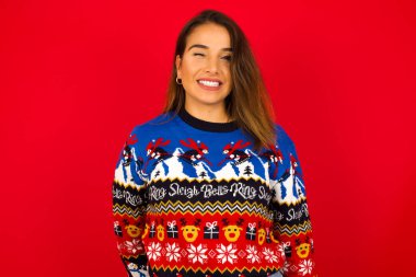 Genç, güzel, beyaz bir kadın, kırmızı duvara karşı Noel süveteri giyiyor, mutlu bir ifade takınarak gözlerini kırpıyor. Yüz ifadeleri ve insanların duygu konsepti.