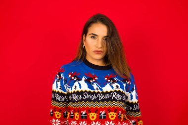 Genç, beyaz bir kadın, kırmızı duvara karşı Noel süveteri giyiyor, kaşlarını çatıyor ve bir şeyden rahatsız oluyor..