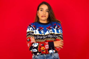 Genç, beyaz bir kadın, kırmızı duvara karşı Noel süveteri giyiyor, ağzını ısırıyor, endişeli ve korkmuş görünüyor..