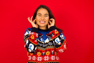Bu sinir bozucu sesi çıkarmayı kes! Mutsuz, genç, güzel, beyaz bir kadın Noel süveterini kırmızı duvara dayamış, endişeli bir yüz ifadesi takınıyor, kulaklarını parmaklarıyla tıkıyor, gürültüden rahatsız oluyor..