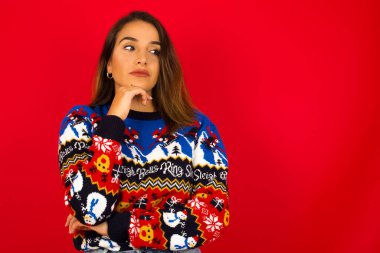 Hayalperest genç beyaz kadın kırmızı duvara karşı Noel süveteri giyiyor, hoş bir ifadeyle, yana bakıyor, elini çenesinin altında tutuyor, hoş bir şeyler düşünüyor..