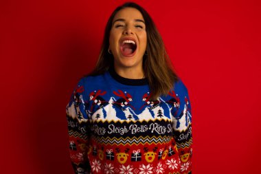 Stresli, genç, beyaz bir kadın Noel süveterini kırmızı duvara dayamış, panik içinde bağırıyor, gözleri dehşet içinde kapanıyor, ellerini başının üstünde tutuyor, dehşet verici haberleri öğreniyor, buna inanamıyorum..