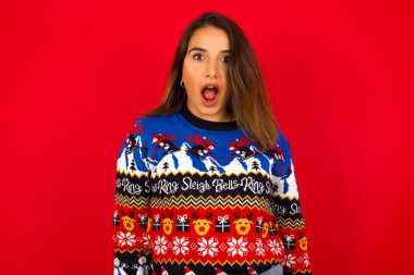 Etkileyici yüz ifadeleri. Şok olmuş genç, beyaz bir kadın Noel süveterini kırmızı duvara dayamış, çenesini kapalı tutuyor gördüklerinden sersemlemiş hissediyor..
