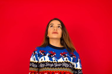Genç, güzel, beyaz bir kadın Noel süveterini kırmızı duvara dayayıp kafasını kaldırıp tuhaf bir şey görüyor..