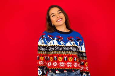 Genç beyaz bir kadın, kırmızı duvara karşı Noel süveteri giyiyor, geniş gülümsemeli, beyaz dişlerini gösteriyor, kendine güvenen bir kadın, izin gününün keyfini çıkarıyor..