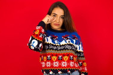 Mutsuz, genç, beyaz bir kadın Noel süveterini kırmızı duvara dayamış, kameraya poz verirken ağlıyor gözyaşlarını eliyle çırpıyor..