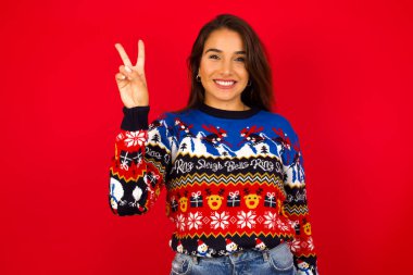 Genç, güzel, beyaz bir kadın Noel süveterini kırmızı duvara dayayıp iki numaralı parmaklarıyla gösterirken kendinden emin ve mutlu bir şekilde gülümsüyor..