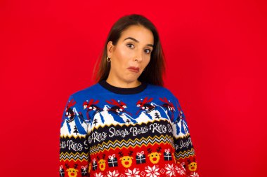 Genç, beyaz bir kadın, kırmızı duvara karşı Noel kazağı giyiyor, züppe bir ifade takınıyor, dudaklarını kıvırıyor ve kaşlarını kaldırıyor, şüpheli ve şüpheci bir ifadeyle bakıyor, şüpheli ve şüpheci..