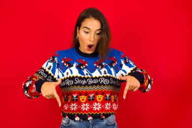 Kırmızı duvara dayalı Noel kazağı giyen beyaz kadın ön parmaklarıyla ağzını açar ve şok geçirir. Reklam kavramı.