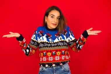 Kafası karışık, beyaz bir kadın Noel süveterini kırmızı duvara karşı giymiş, omuzlarını silkip, 