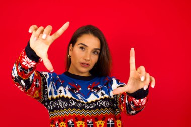 Kırmızı duvara karşı Noel süveteri giymiş gülümseyen beyaz kadın portresi. Kameraya bakıyor ve el hareketi yapıyor. Yaratıcılık ve fotoğrafçılık kavramı.