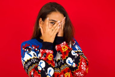 Beyaz kadın Noel süveteri giyip kırmızı duvara yaslanmış elleriyle yüzünü kapatıyor ve bir gözü parmaklarının arasında dışarı bakıyor. Bir şeyden ya da birinden korktun mu?.