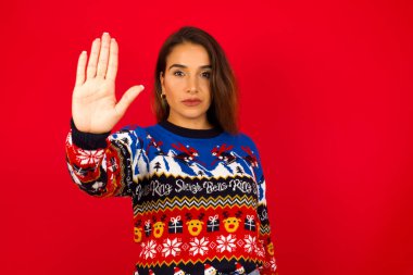 Beyaz kadın kırmızı duvara karşı Noel süveteri giyip avuç içiyle el hareketi yapıyor. Yüzünde olumsuz ve ciddi bir jest olan uyarı ifadesi.