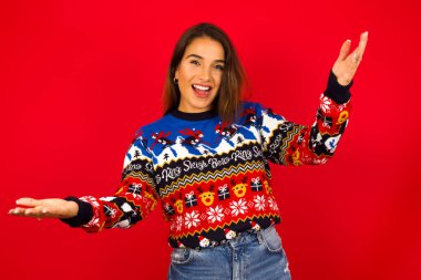 Kırmızı duvara karşı Noel süveteri giyen neşeli beyaz kadın hoş geldin jesti yapıyor ve kollarını başının üstüne kaldırıyor..