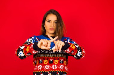 Kırmızı duvara karşı Noel süveteri giyen beyaz bir kadın olumsuz işaret veren kızgın bir ifadeye sahip..