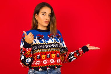 Kırmızı duvara karşı Noel süveteri giyen neşeli beyaz kadın başparmağını kaldırıp diğer elini işaret ediyor. Bunu tavsiye ederim..