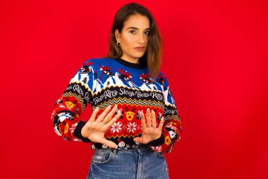 Kırmızı duvara karşı Noel kazağı giyen beyaz bir kadın korkmuş bir ifade takınıyor ve iki eliyle birden 