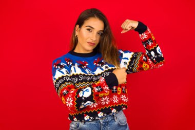 Kırmızı duvara karşı Noel kazağı giyen, gülümseyen beyaz kadın kaslarını göstermek için elini kaldırıyor, zaferden emin, güçlü ve bağımsız..