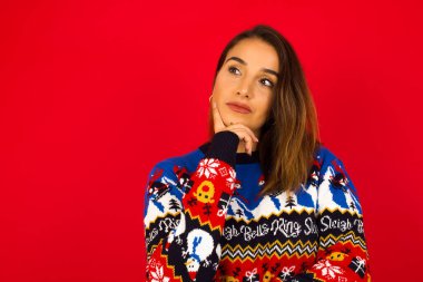 Yüz ifadeleri ve duygular. Kafkasyalı düşünceli bir kadın Noel süveteri giyip kırmızı duvara yaslanmış kafasının altında el ele tutuşmuş, şüpheli bakıyor..