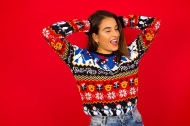 Beyaz kadın Noel süveteri giyiyor kırmızı duvara yaslanmış gevşiyor ve esniyor, kolları ve elleri başının arkasında mutlu bir şekilde gülümsüyor.