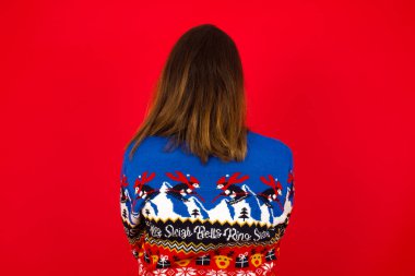 Beyaz kadın, kırmızı duvara karşı Noel süveteri giyiyor. Arkaya bakıyor. Kolları vücudunda..