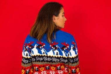 Kırmızı duvara karşı Noel süveteri giyen beyaz bir kadının arka görüntüsü.