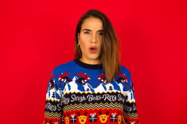 Kafkasyalı bir kadın kırmızı duvara karşı Noel kazağı giyiyor. Tiksinti, isteksizlik, gergin bakışlar, somurtkan bakışlar, bir şeye kızgın bakışlar..