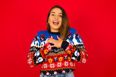 Kırmızı duvara karşı Noel süveteri giyen mutlu beyaz kadının elleri göğsünde. İnsani duygular, gerçek duygular ve yüz ifadesi kavramı.