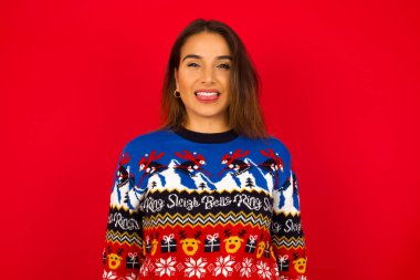 Kırmızı duvara karşı Noel süveteri giyen, gülümseyen ve dil gösteren beyaz bir kadın..
