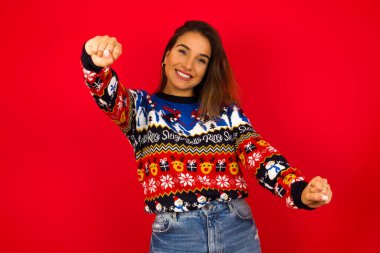 Kırmızı duvara karşı Noel süveteri giyen beyaz bir kadının portresi, ellerini başının üstünde tutarken geniş bir gülümseme. Güven ve rahatlama kavramı.