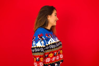 Sağlıklı ciltli kırmızı duvara karşı Noel süveteri giyen beyaz kadın profili, derin düşüncelere dalmış, açık havada yürümeye hazır..