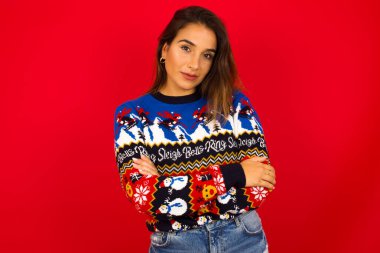 Kırmızı duvara karşı Noel süveteri giyen, kendine güveni yüksek, kollarını kavuşturan, meslektaşlarıyla tanıştığı için mutlu olan beyaz bir kadının belden yukarı pozu..