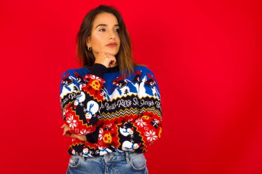 Kafkasyalı bir kadın kırmızı duvara karşı Noel kazağı giyiyor. Çenesinin altında elleri ve yana bakıyor. Şüpheli ve şüpheci bir ifadeyle..  