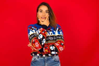 Kızıl duvara karşı Noel süveteri giyen genç, güzel beyaz bir kadın. Ağzı ellerle kapatıyor. Bir şeyden ya da birinden tırnak yemekten korkuyor.