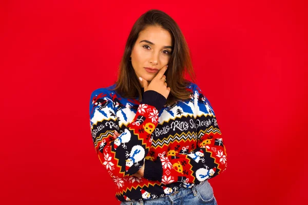 Kırmızı duvara karşı Noel süveteri giyen düşünceli beyaz kadın elini çenesinin altında tutuyor, doğrudan kameraya bakıyor ve ilgi çekici bir şey dinliyor. Gençlik kavramı.