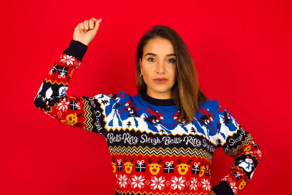 Genç, beyaz bir kadın, kırmızı duvara karşı Noel kazağı giyiyor, ciddi, güçlü ve isyankar hissediyor, yumruğunu kaldırıyor, politik devrim için protesto ediyor ya da savaşıyor..