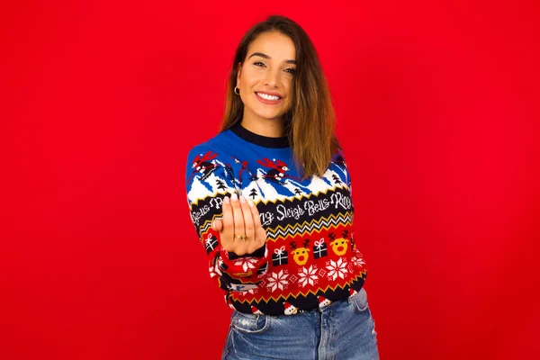 年轻美丽的白种人女人 身穿圣诞毛衣 靠着红墙 邀请你来 带着自信和微笑用手做手势 积极友善 — 图库照片