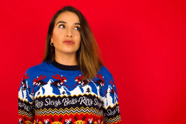 Genç, beyaz bir kadın, kırmızı duvara karşı Noel kazağı giyiyor, düşünceli bir şekilde kenara bakıyor, üniversiteden sonra planlar yapıyor, yeni proje hakkında ne yapacağını düşünüyor..